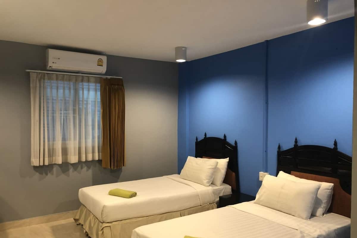 A hotel room with two single beds, a window with curtains, an air conditioning unit, and a blue accent wall.