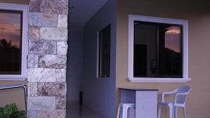 A covered outdoor area with a stone pillar, a small desk, two plastic chairs, and a window with a sunset view. - Heilee's Guest House (General Santos)
