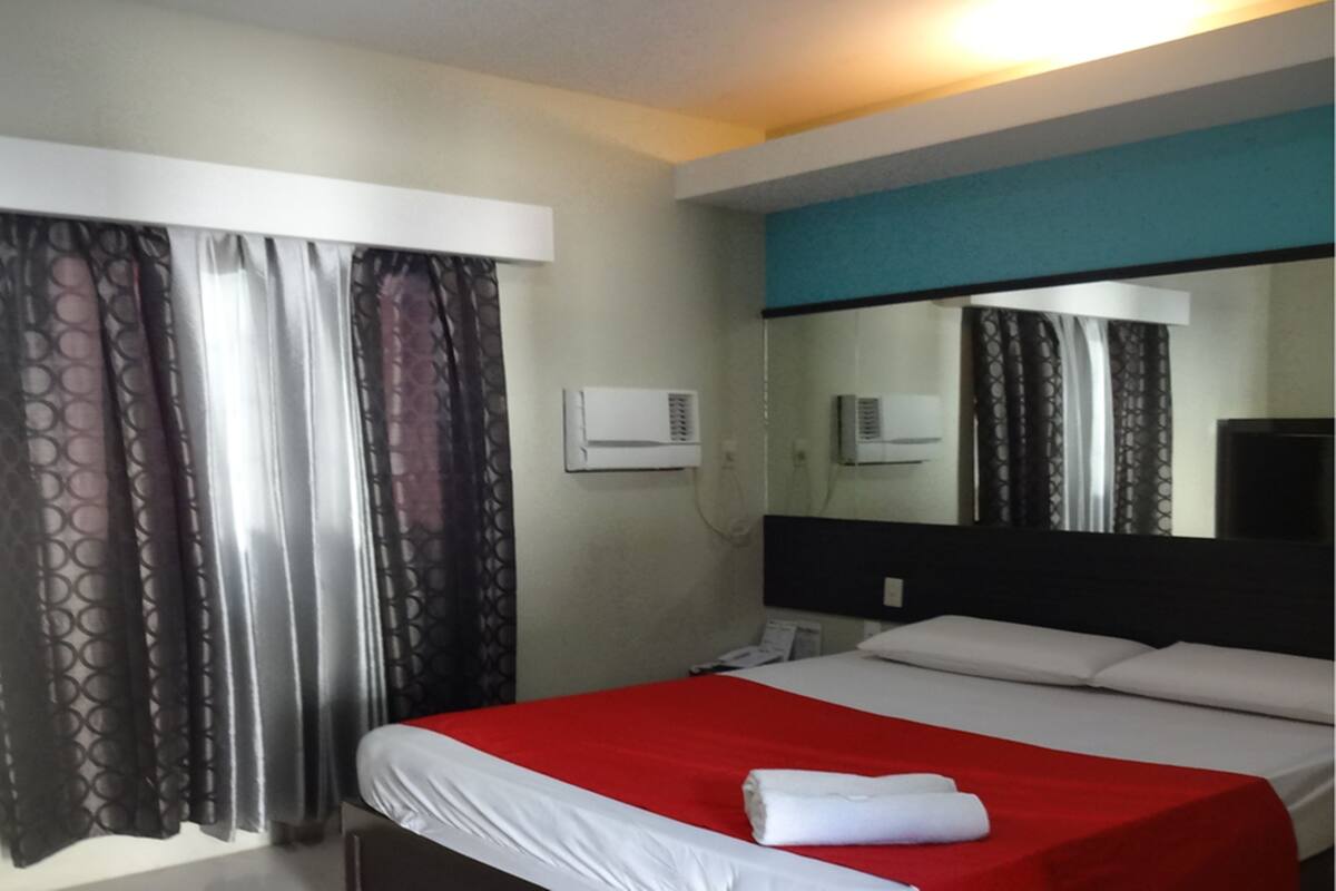 A hotel room with a bed, curtains, an air conditioning unit, and a mirror.