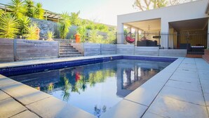A modern outdoor swimming pool with a clear blue edge, surrounded by a stone wall and lush greenery. - Hollywood Tree House (Burnside)