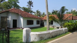 A white building with a red-tiled roof, surrounded by a well-maintained lawn and palm trees. - Mendis Cottage (Ahangama)