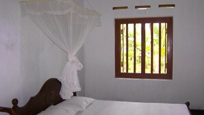 A single bed with white bedding, a wooden headboard, and a mosquito net. There is a window with vertical bars and a view of greenery outside. - Mendis Cottage (Ahangama)