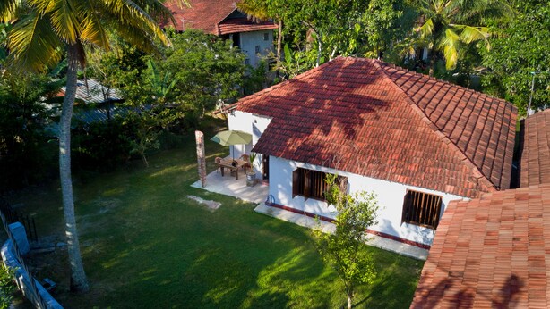 A white house with a red-tiled roof, surrounded by lush greenery and palm trees. - Mendis Cottage (Ahangama)