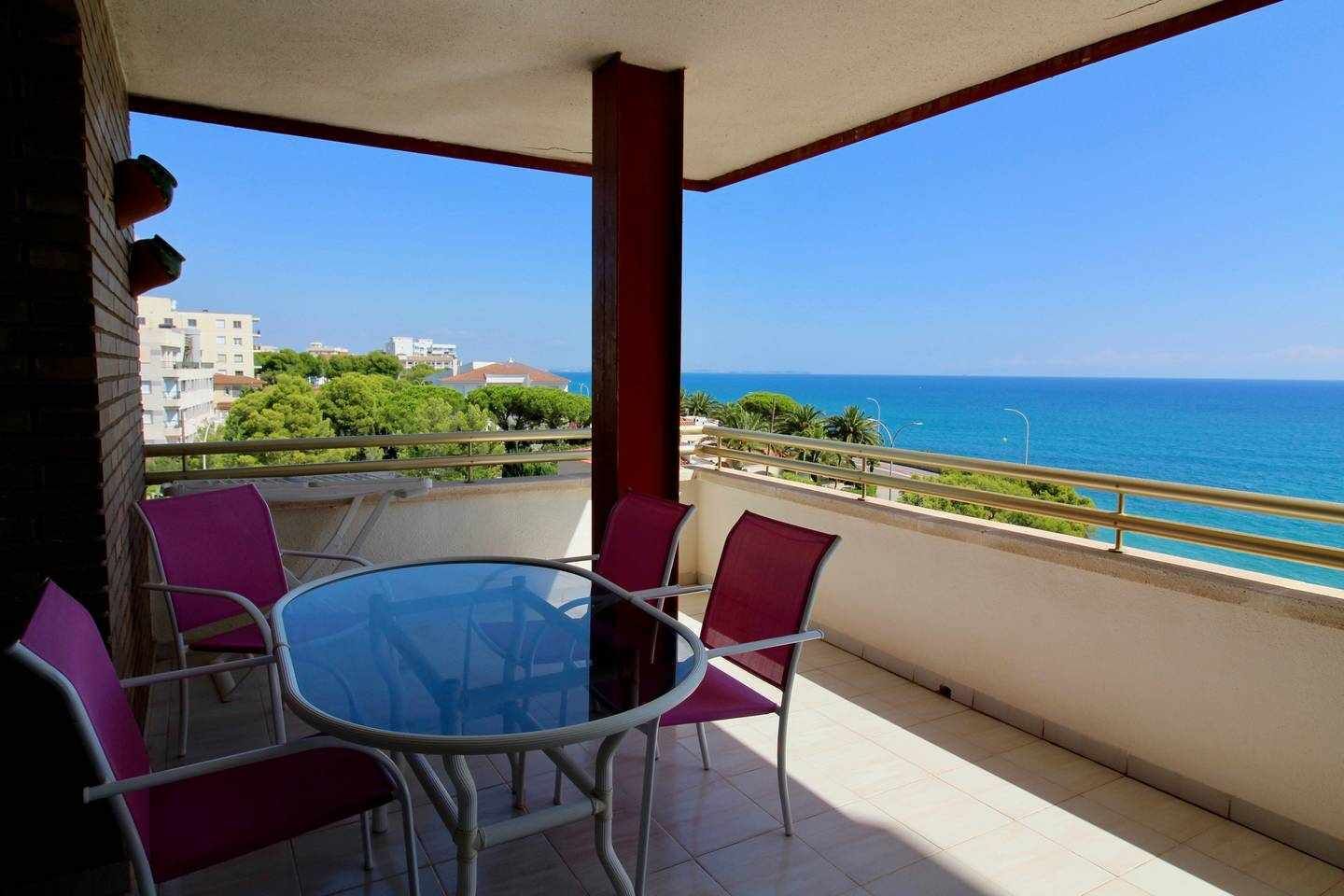 Un balcón con una mesa redonda de cristal y cuatro sillas, con vistas a la costa, edificios y follaje al fondo.
