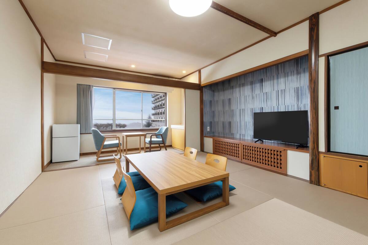 Une chambre d’hôtel moderne avec une table à manger en bois, des chaises rembourrées bleues, un téléviseur à écran plat et une grande fenêtre donnant sur la ville.