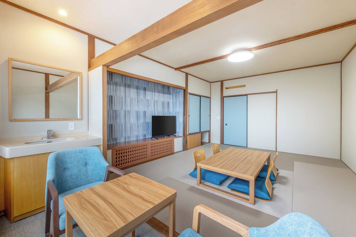 Une pièce comprenant une table à manger en bois, des chaises bleues, un téléviseur posé sur un meuble en bois, un petit lavabo et un miroir.
