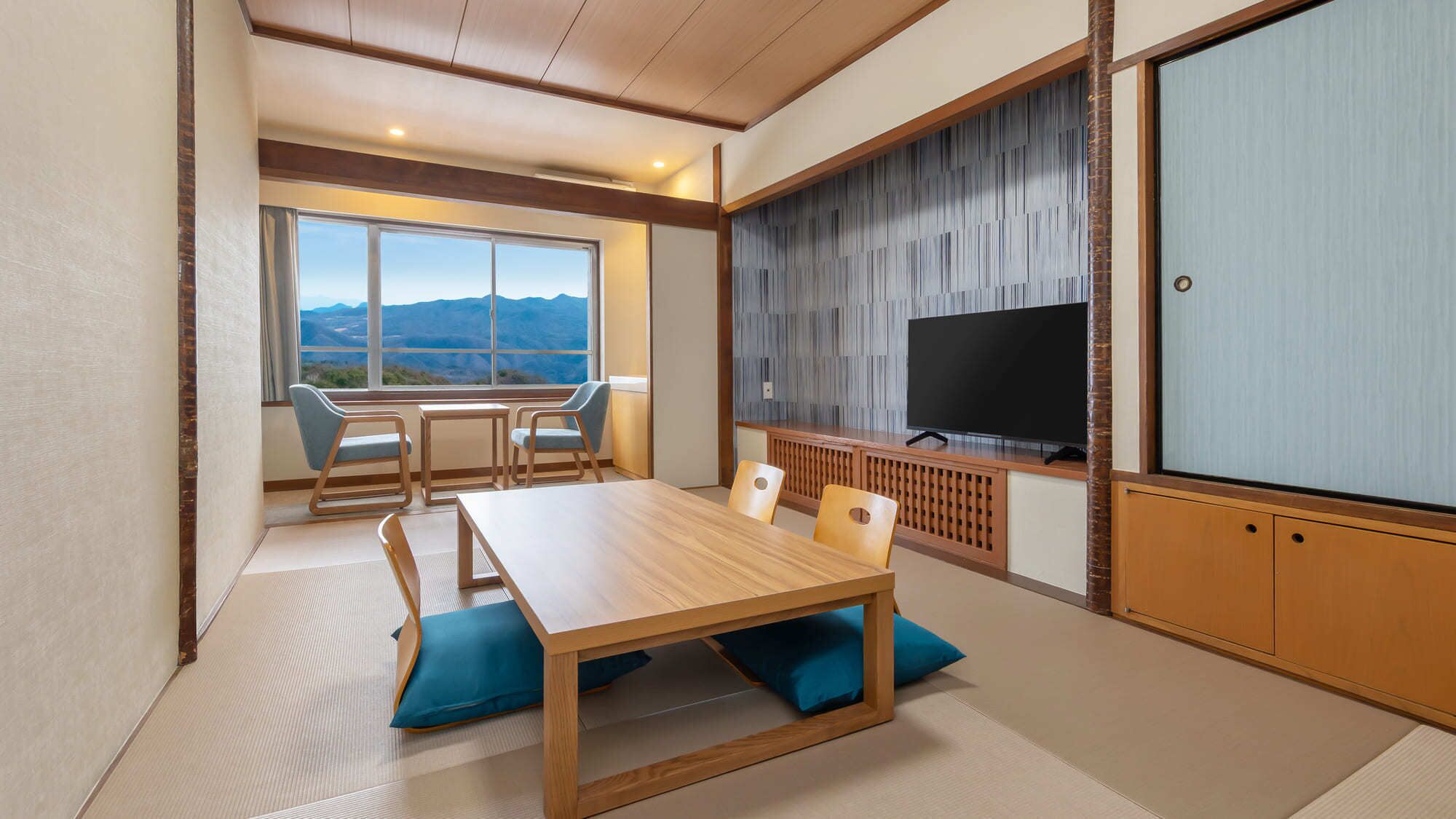 A room with a wooden dining table, chairs, a television, and a view of mountains through large windows.