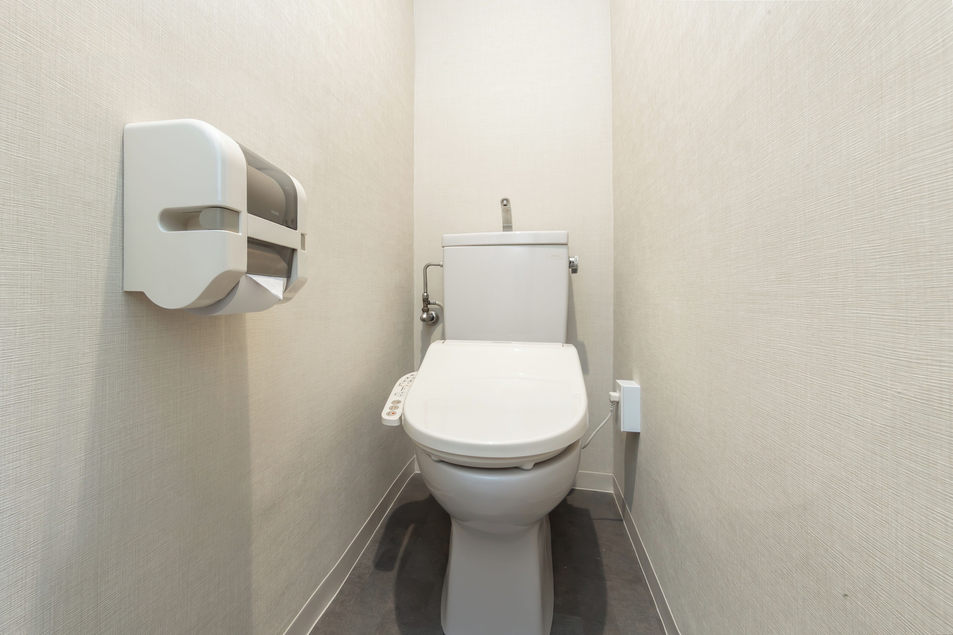 Une petite salle de bain moderne, équipée de toilettes, d’un distributeur de papier fixé au mur et d’un mur sobre.
