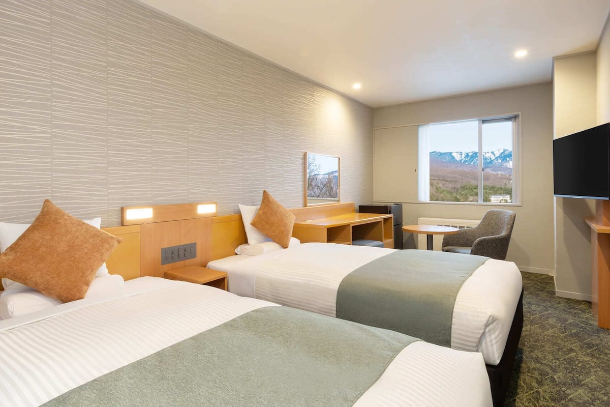 Habitación de hotel con dos camas, un escritorio, una silla, un televisor y ventana con vista a la montaña.