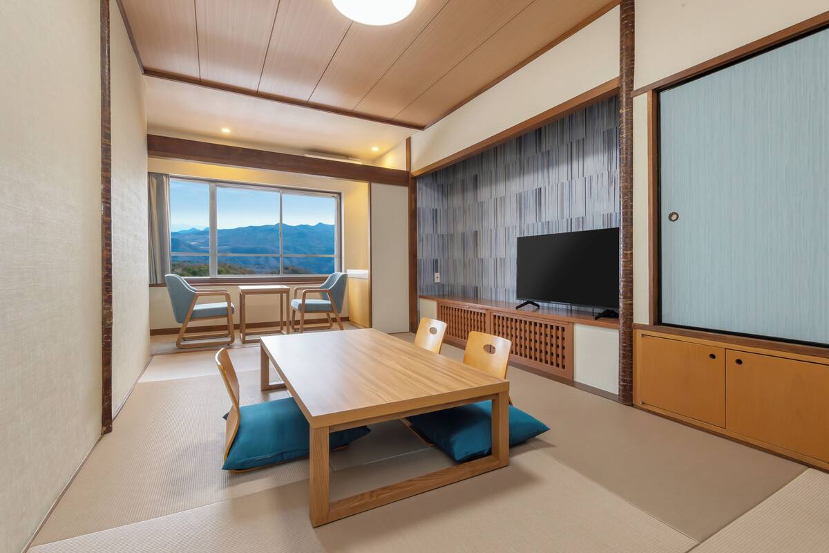 Une pièce avec un sol recouvert de tatamis, une table en bois, des chaises, un téléviseur et une vue sur les montagnes.