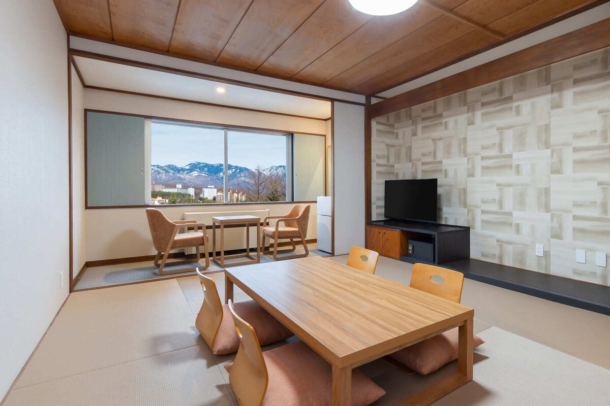 A modern living room with a wooden dining table, chairs, a TV, and a view of mountains through large windows.