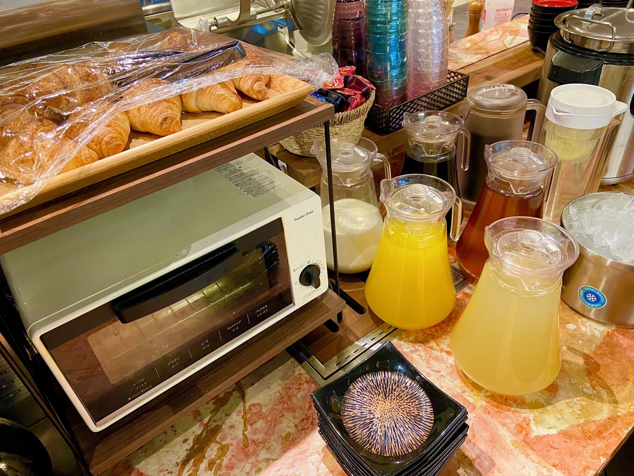 A kitchen counter with a toaster oven, plastic-wrapped pastries, and various beverage containers.