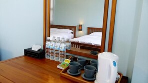 A hotel room with a wooden table, a bed with white linens, a mirror, and various amenities including bottled water, mugs, and a kettle.