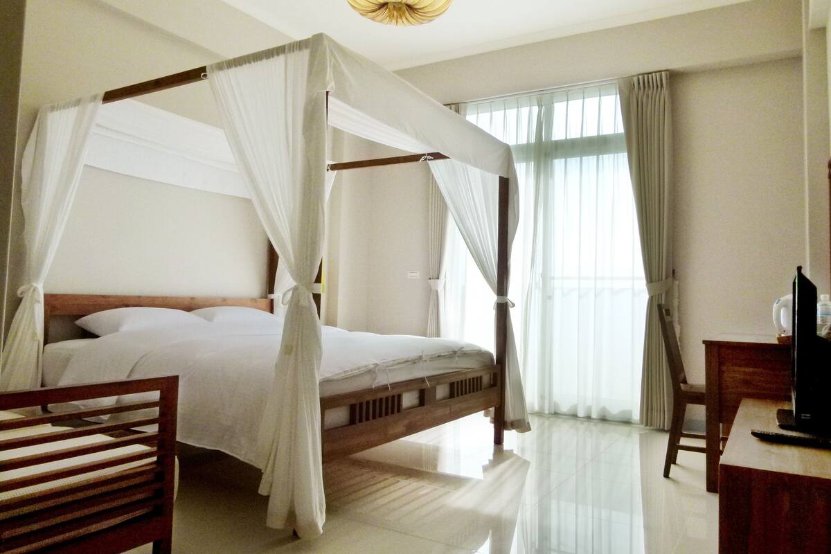 A four-poster bed with a canopy, a wooden desk, and a chair in a hotel room with large windows and a chandelier.