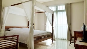 A four-poster bed with a canopy, a wooden desk, and a chair in a hotel room with large windows and a chandelier.