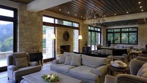 A spacious living room with a stone fireplace, comfortable seating, and large windows offering a view of the outdoors. - Styga Mountain Resort (Aigialeia)