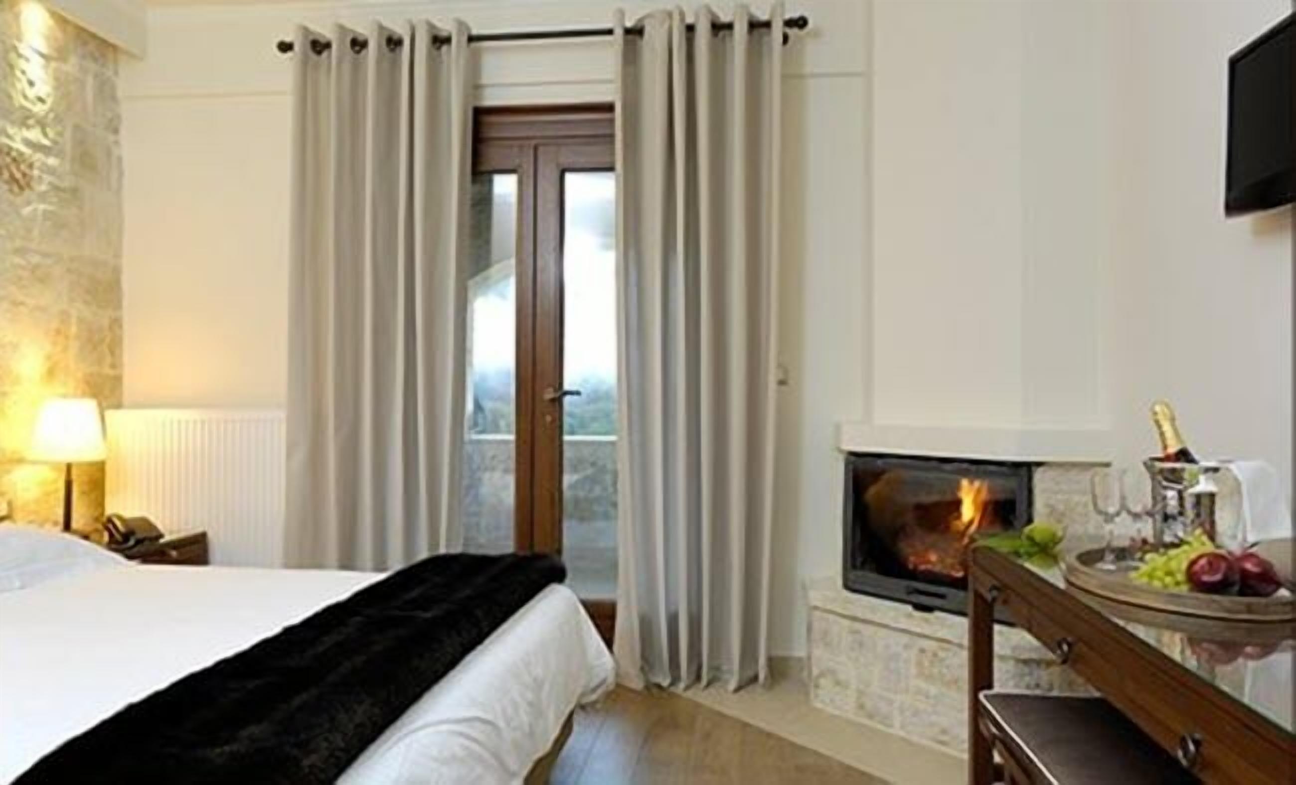 A hotel room with a bed, a fireplace, a view through the window, and a table with a bottle and fruits.