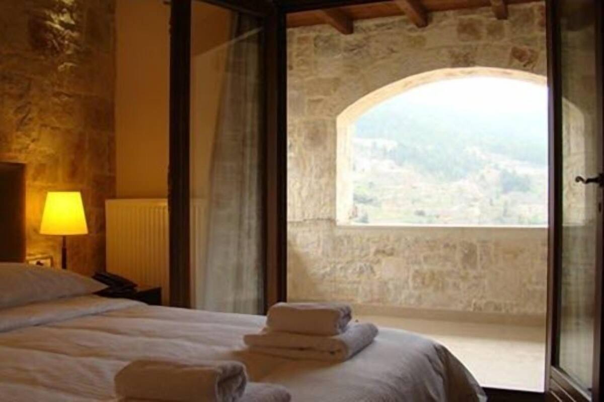 A bedroom with a stone wall, a bed with white linens, a nightstand with a lamp, and a view through an arched window.