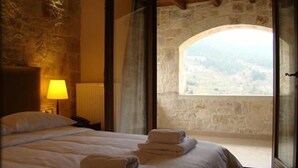 A bedroom with a stone wall, a bed with white linens, a nightstand with a lamp, and a view through an arched window. - Styga Mountain Resort (Aigialeia)