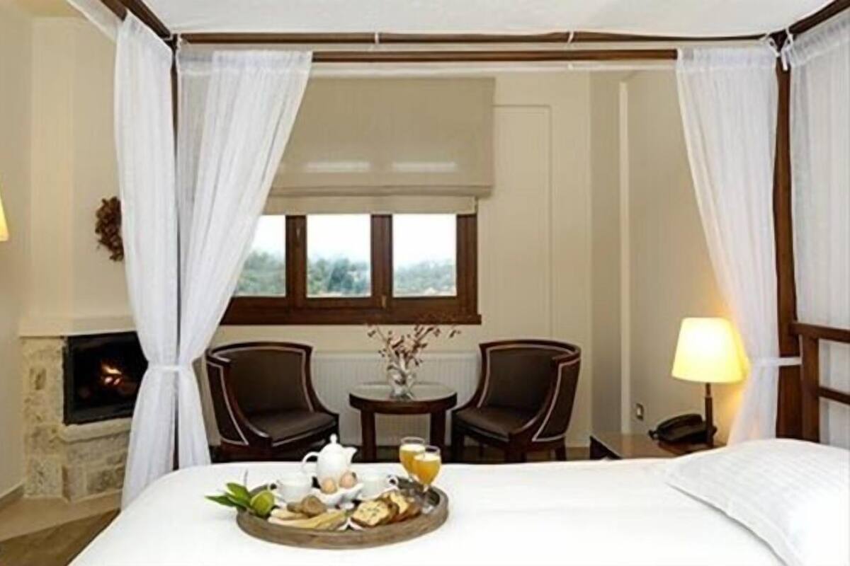 A bedroom with a four-poster bed, a fireplace, two chairs, a small table with a vase, and a tray of breakfast items.