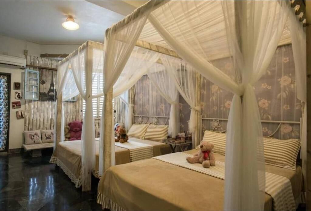 Une chambre avec quatre lits, chacun surmonté d’un dais. Des peluches sont posées sur les lits, et la chambre présente un décor classique avec un papier peint à motifs floraux.