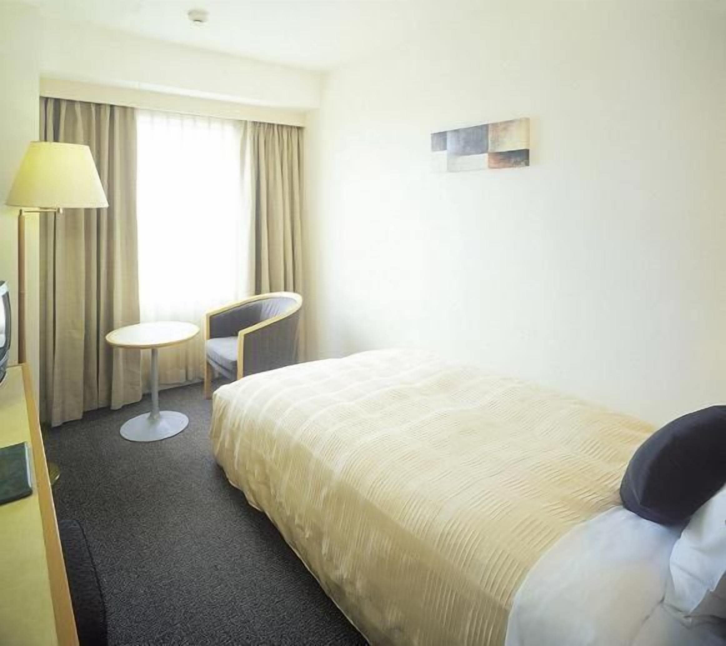 A hotel room with a bed, a desk, a chair, a lamp, and a window with curtains.