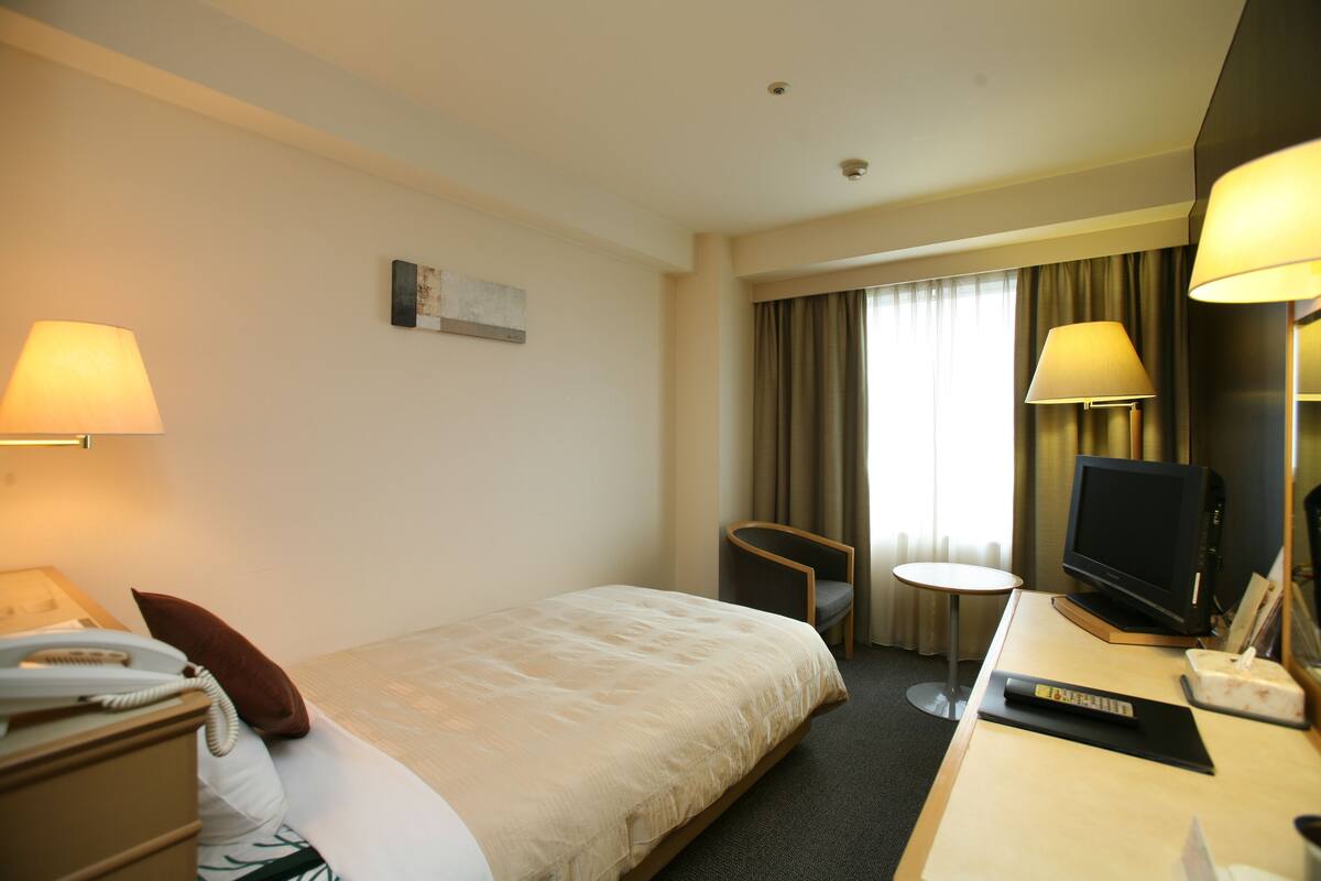 Een hotelkamer met een bed, nachtkastje, lamp, stoel, televisie en een klein tafeltje.