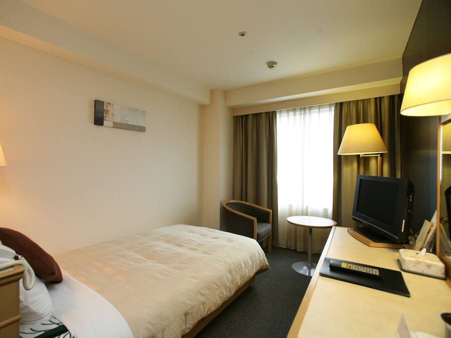 Quarto de hotel com cama, mesa de cabeceira, abajur, cadeira, televisão e uma pequena mesa.