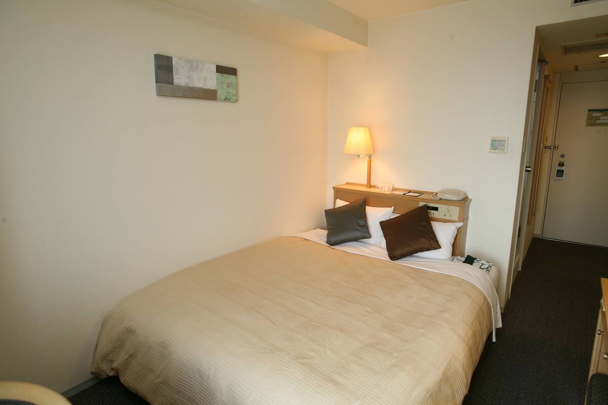 Een hotelkamer met een groot bed, een nachtkastje met een lamp en een deur naar een andere kamer.