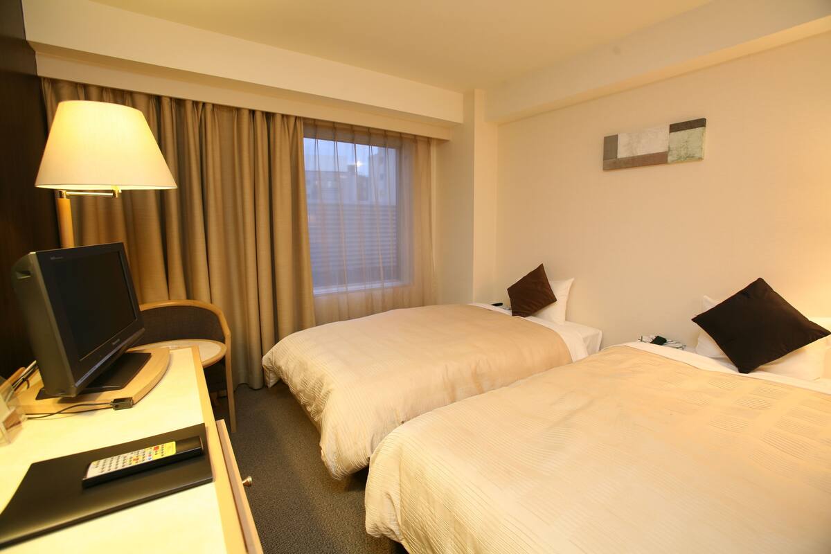 Een hotelkamer met twee bedden, een bureau met een computer, een lamp en een raam met gordijnen.