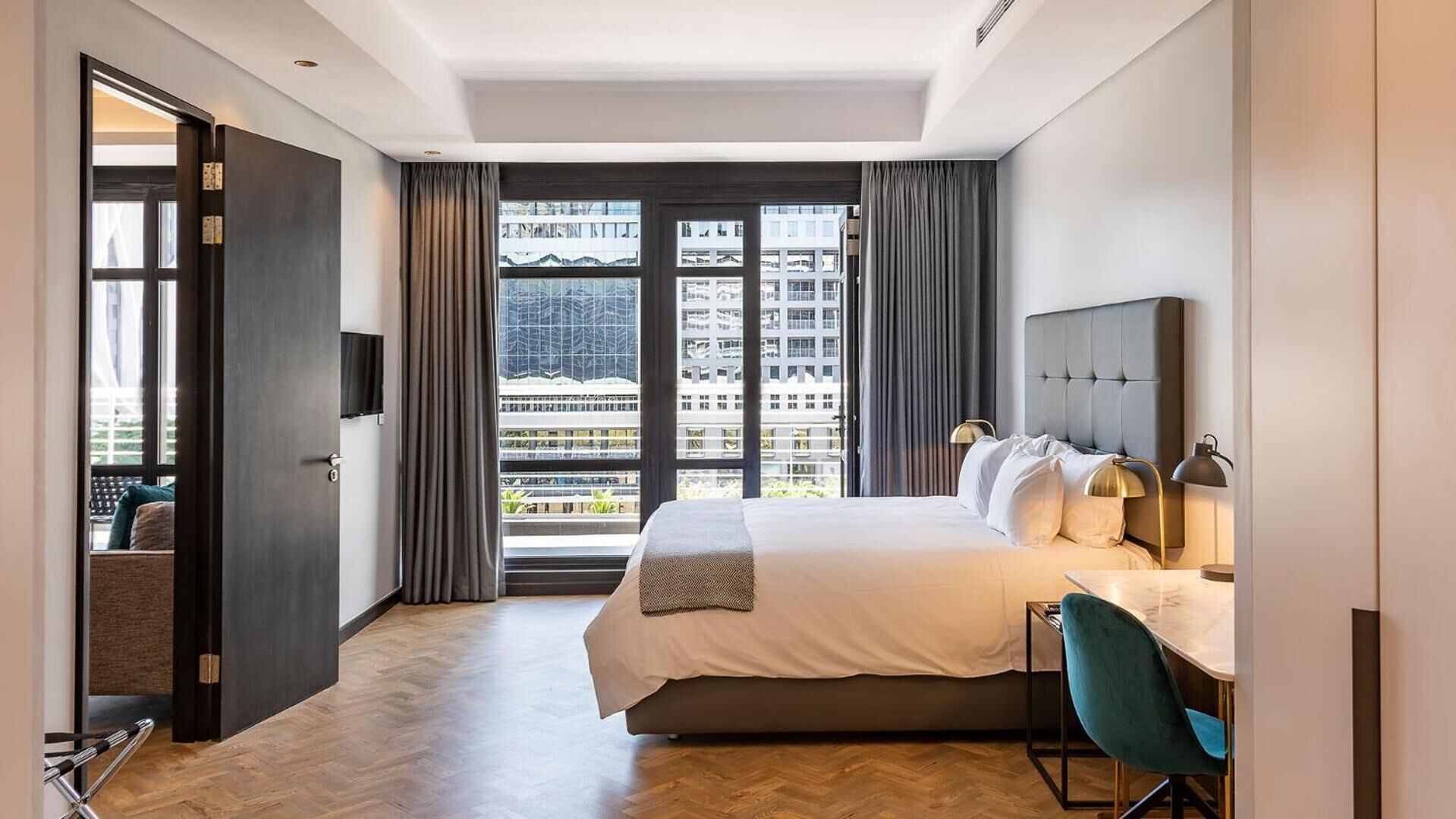 A modern hotel room with a large bed, a desk with a chair, a TV, and a view of buildings through large windows.