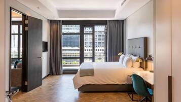 A modern hotel room with a large bed, a desk with a chair, a TV, and a view of buildings through large windows.