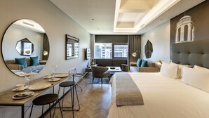 Una habitación de hotel moderna con una cama grande, una zona de comedor con mesa y sillas, un sofá y un espejo amplio.
