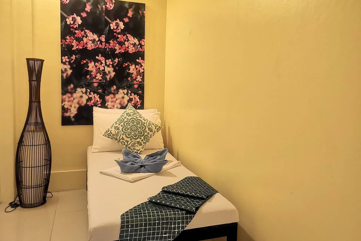 A small, neatly made bed with a pillow and a folded towel, a black decorative vase, and a floral painting on the wall.