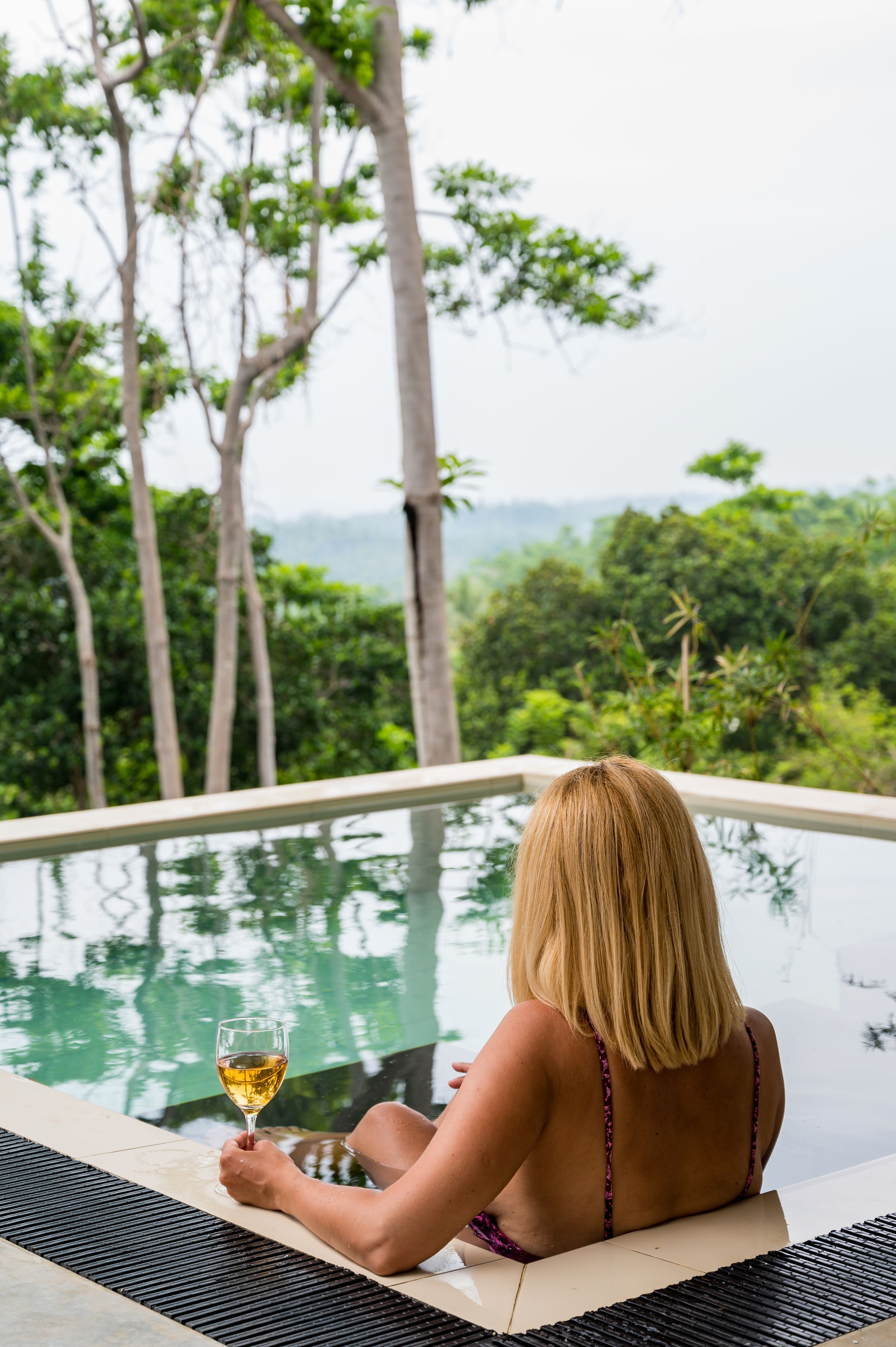 Una persona sentada junto a una piscina infinita, con una copa de vino, rodeada de exuberante vegetación y árboles.
