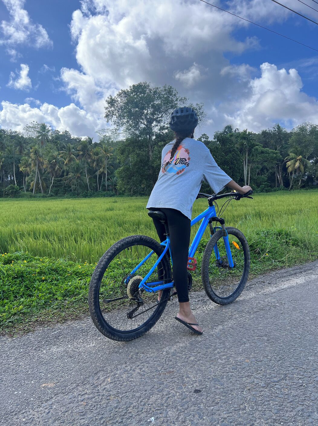 A person riding a blue bicycle on a rural road with green fields and trees in the background.