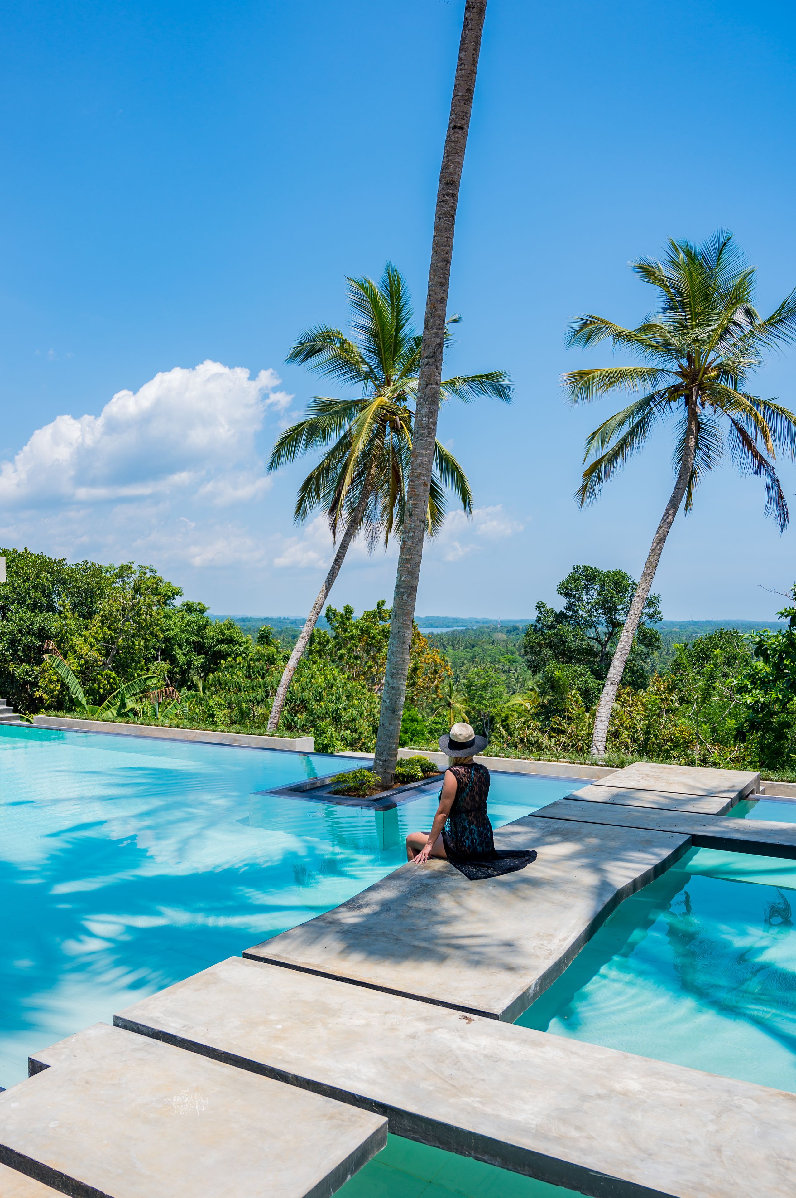 A pool with clear blue water, surrounded by palm trees and lush greenery, with a person sitting on the edge.