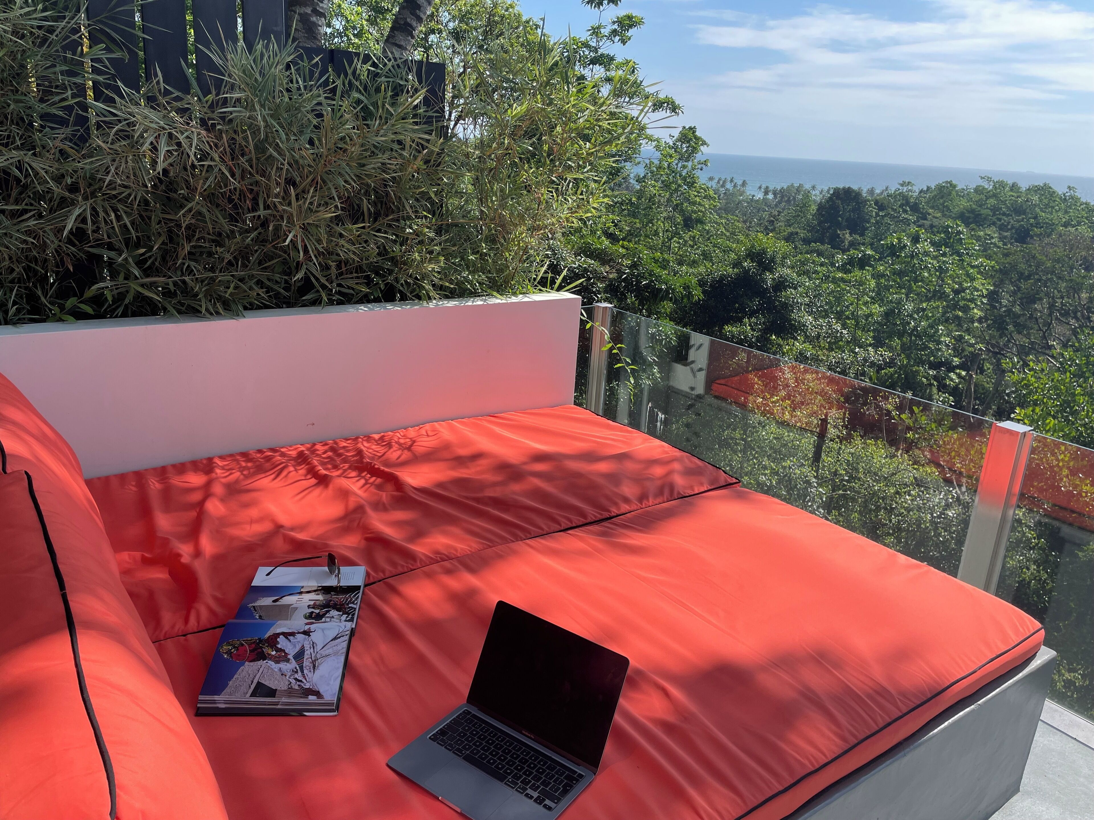 A red cushioned seating area with a laptop and a magazine, overlooking a lush landscape and the sea.