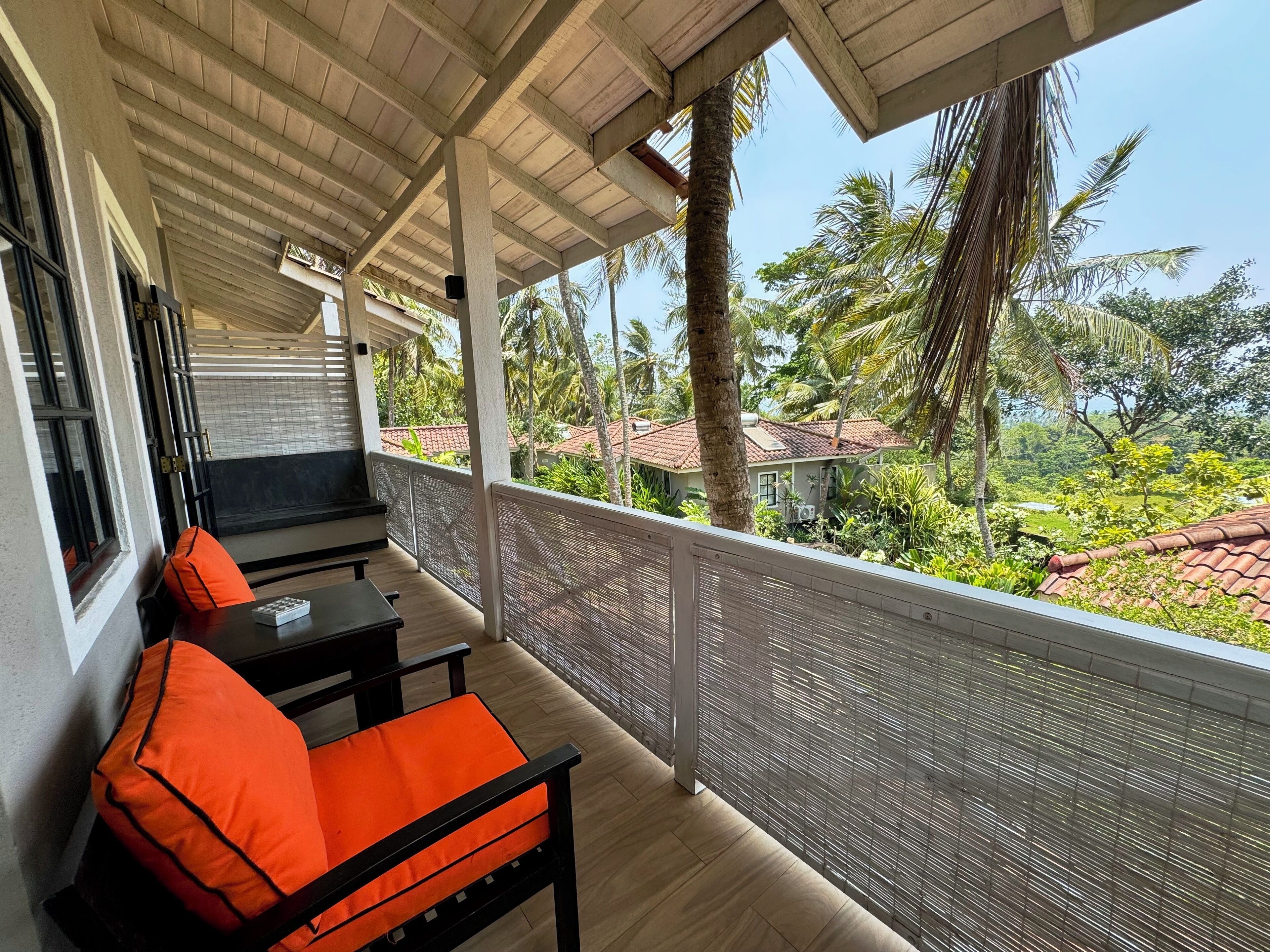 A covered balcony with wooden flooring, a black table, and orange chairs.