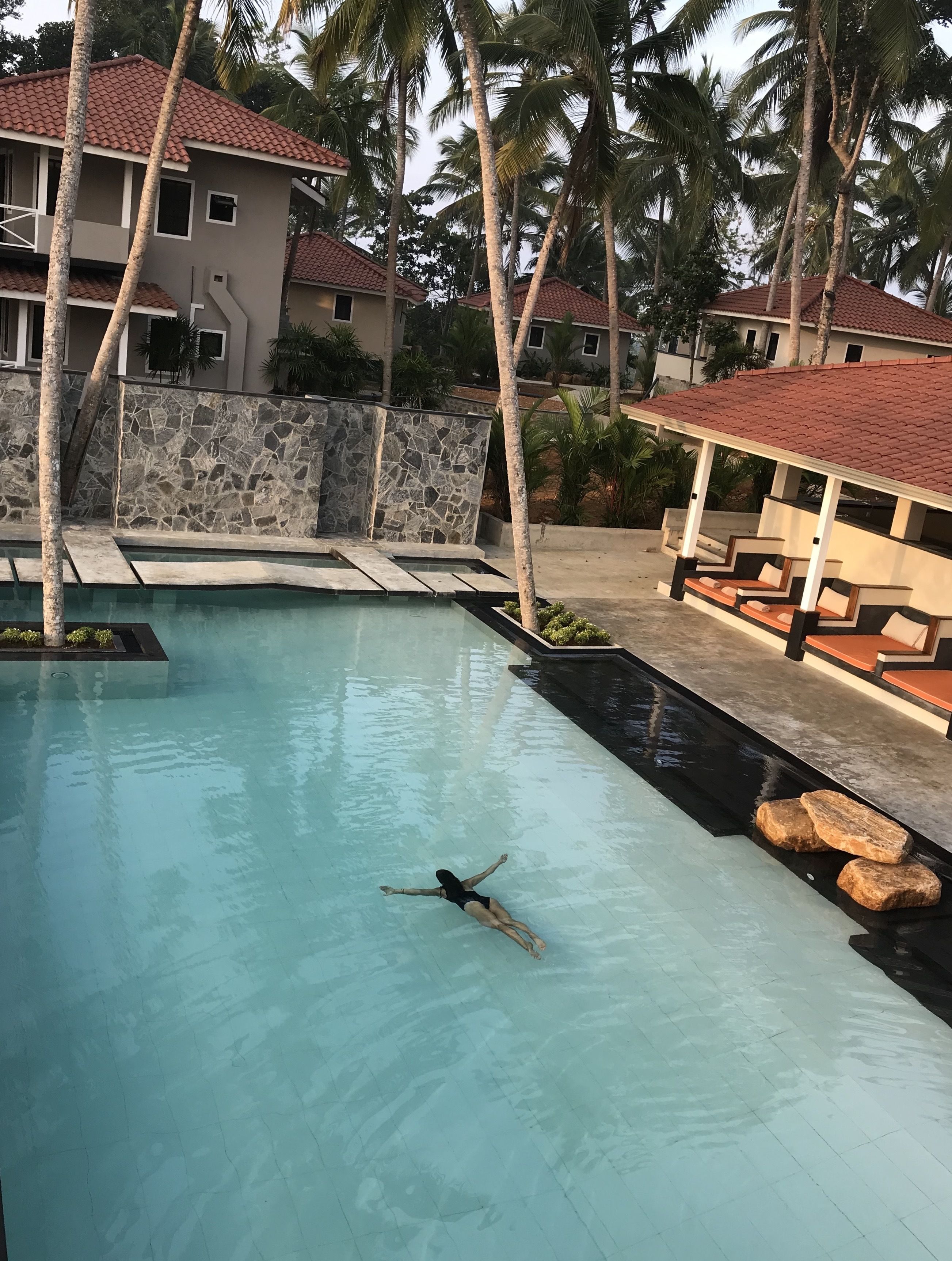 Ein Schwimmbecken mit einer schwimmenden Person, umgeben von Palmen und einer Steinmauer.