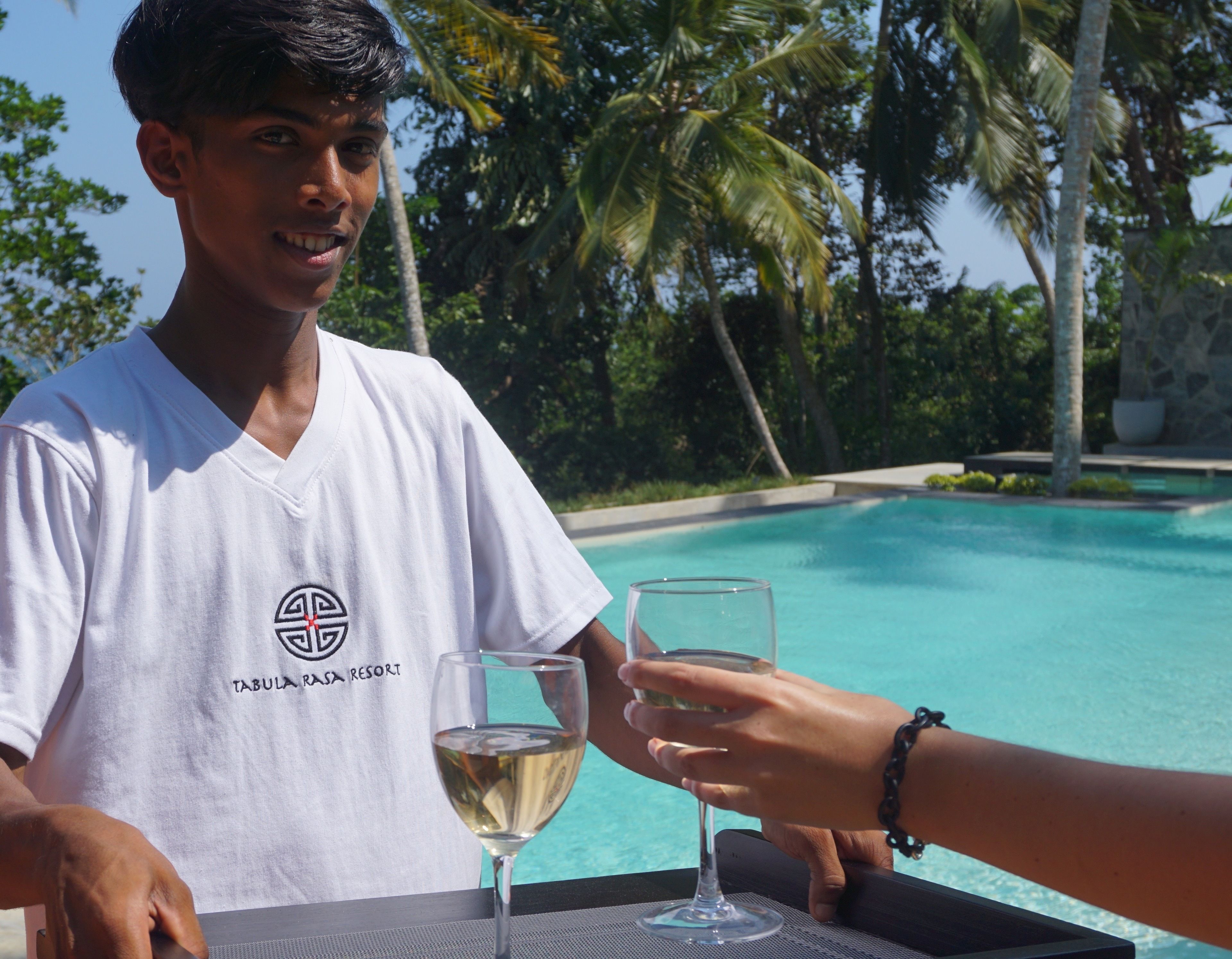 A person in a white T-shirt with a logo, holding a glass of white wine, standing by a pool with palm trees in the background.