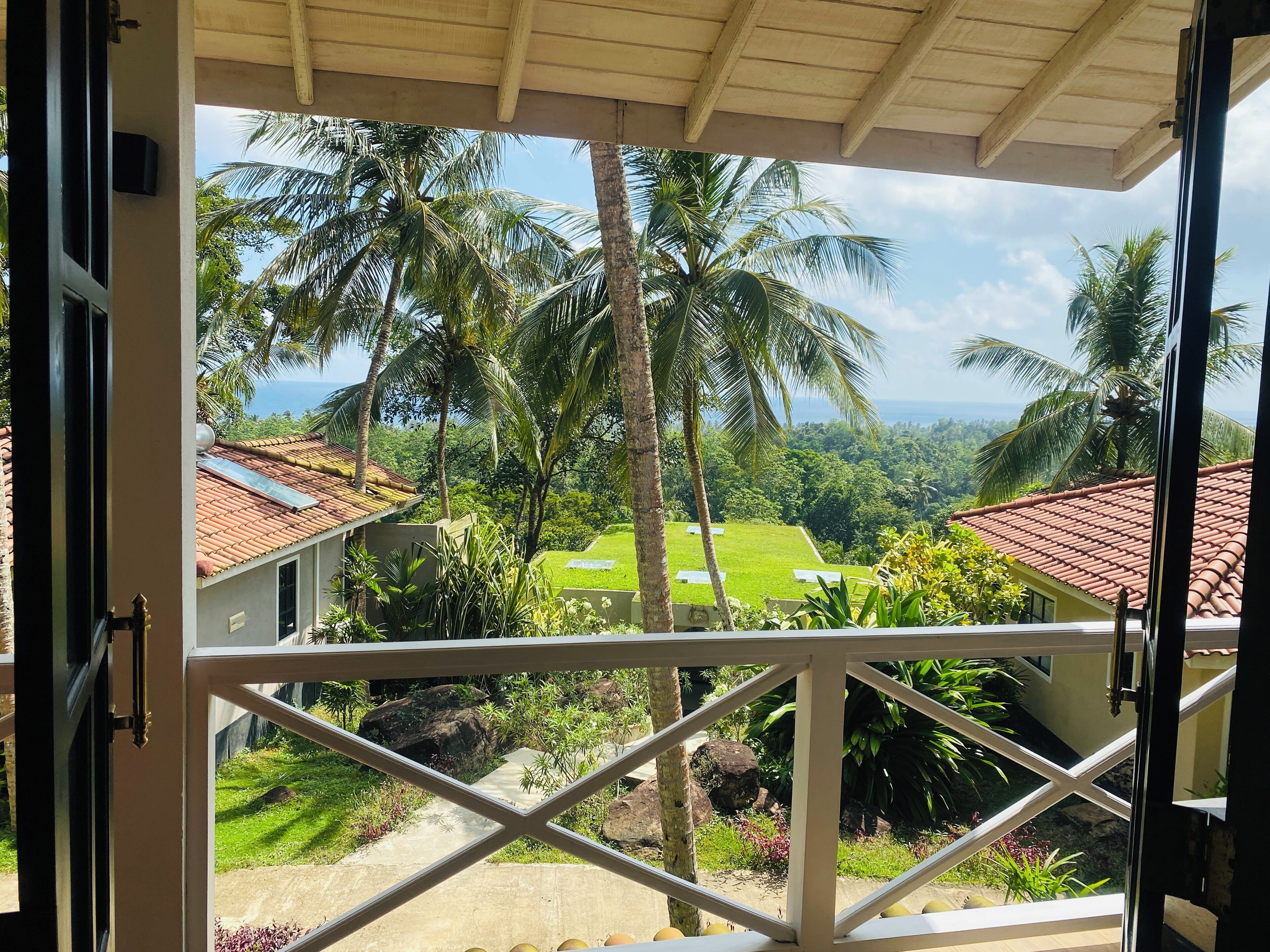 A view from a balcony with a tiled roof, overlooking a green lawn, palm trees, and a distant view of the ocean.