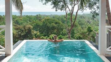Ein rechteckiger Infinity-Pool mit klarem, blauem Wasser, umgeben von weißen Säulen und üppiger Vegetation, mit Blick auf einen dichten Wald und den Ozean.