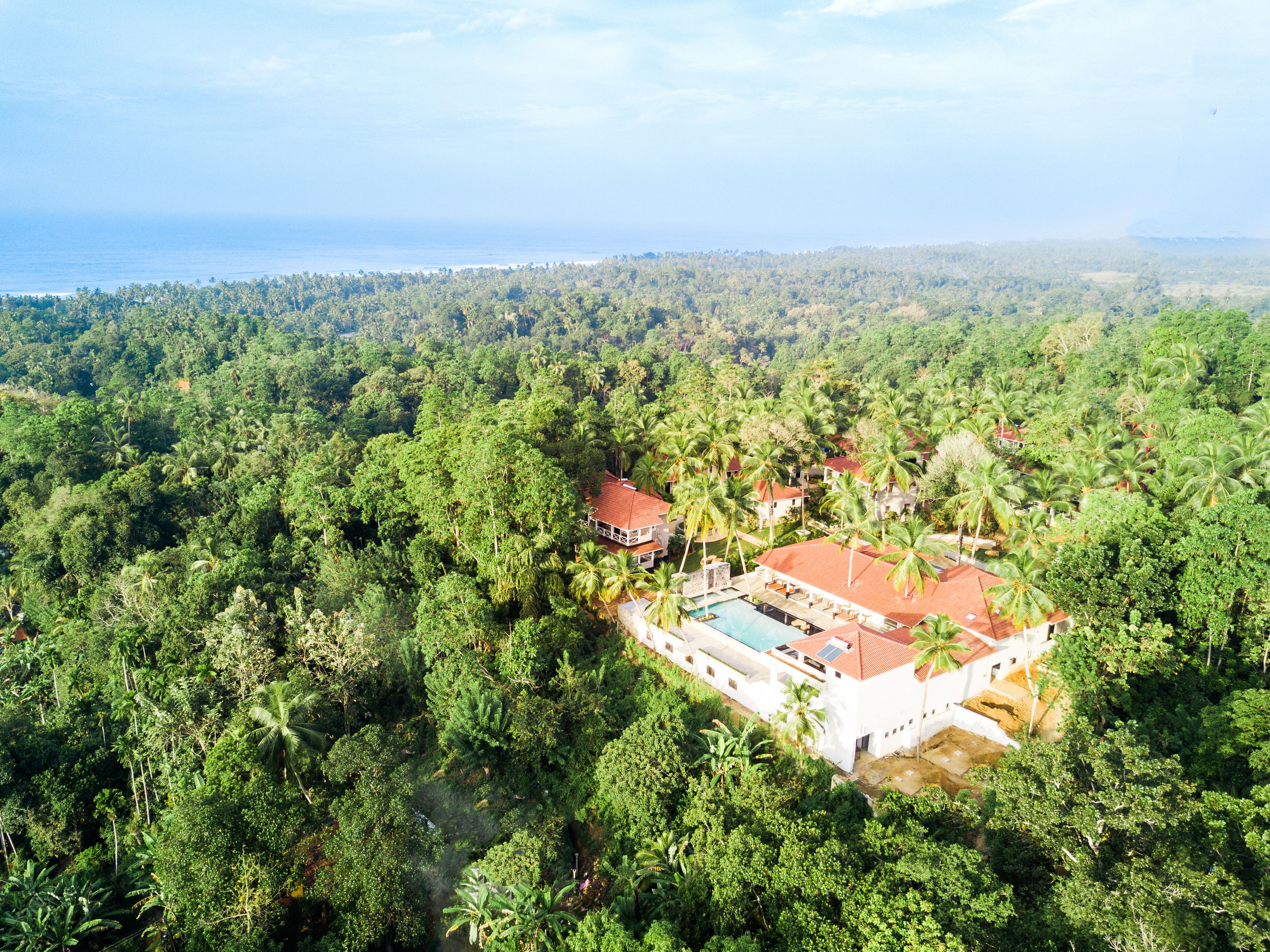 An aerial view of a resort with a swimming pool, surrounded by dense tropical vegetation and a view of the ocean.