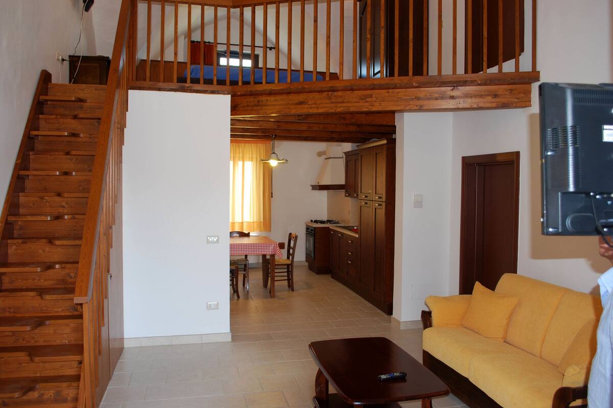 Uma sala de estar ampla com escada de madeira, mesa de jantar com cadeiras, sofá e área de cozinha com armários de madeira e televisão.