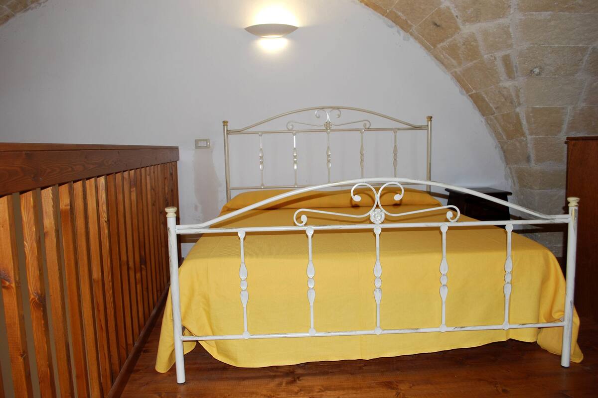Uma cama de solteiro com estrutura de metal e colcha amarela em quarto com paredes de pedra e um banco de madeira.