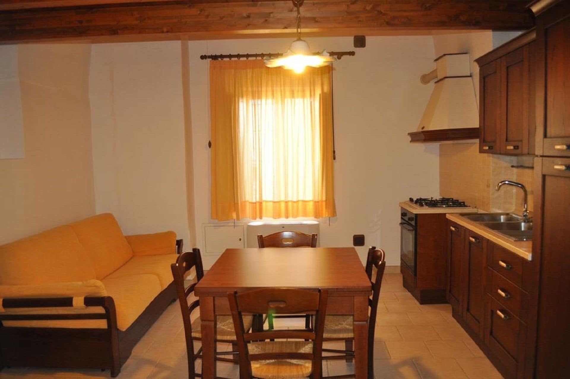A compact kitchen with wooden cabinets, a dining table with chairs, and a window with curtains.