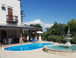Une piscine avec une fontaine, un patio couvert avec des chaises et un bâtiment doté d’un balcon.