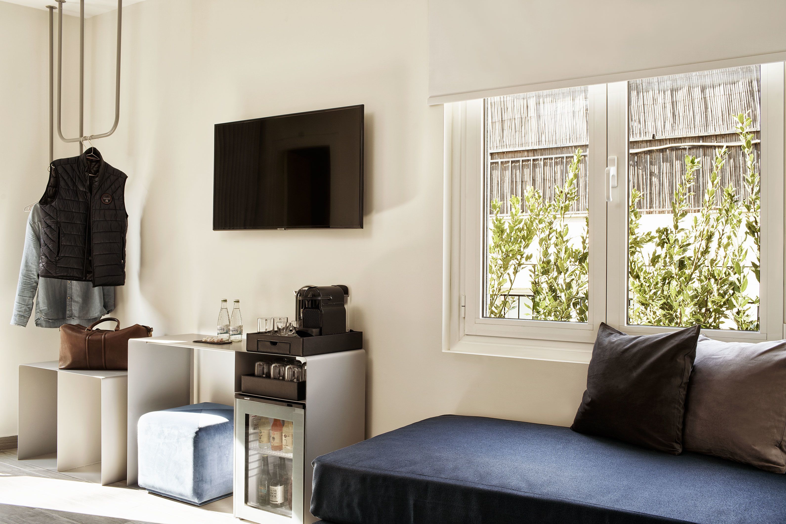 Quarto moderno com uma cama, TV fixada na parede, cafeteira e janela com vista para o verde.