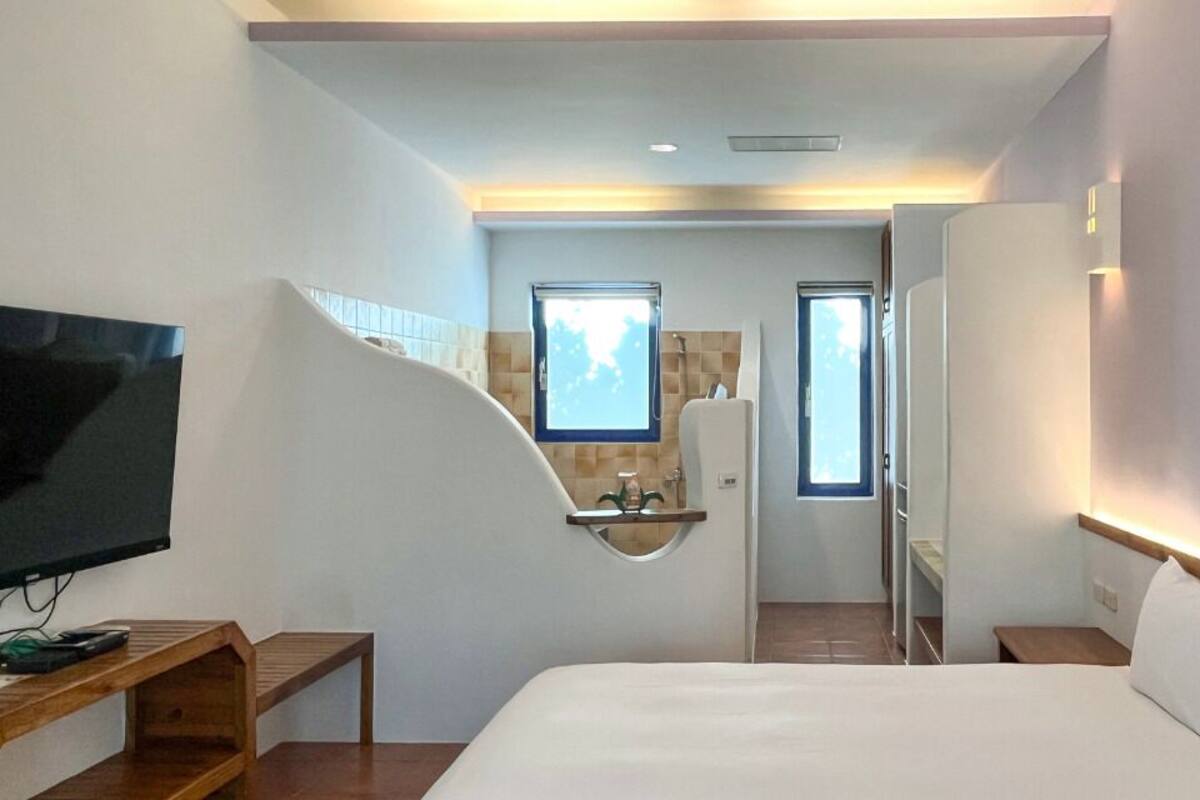 A hotel room with a bed, a TV, a wooden bench, a bathroom with a mirror, and a window.